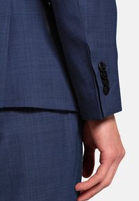 Nahaufnahme einer Hand, die seitlich ruht, mit einem blauen Anzugjackett und passender Hose, mit schwarzen Knöpfen an den Ärmeln.