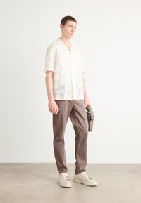 Lichte, doorzichtige kortweg shirt met een subtiel patroon, beige broek en crèmekleurige sneakers. Het model houdt een kleine bruine tas vast.