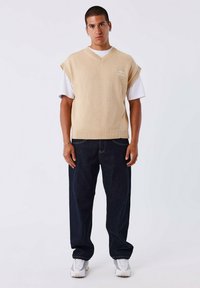 Suéter sin mangas de punto beige con escote en V, con bordes acanalados, llevado sobre una camiseta blanca, combinado con jeans de pierna ancha azul oscuro y zapatillas blancas.