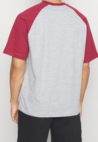 Camiseta gris de manga corta con mangas raglán rojas, cuello redondo y textura suave. La parte posterior presenta un diseño liso, sin adornos.