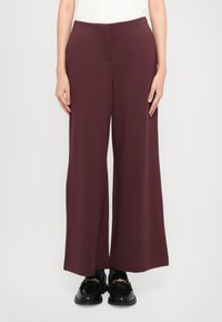 Pantalons larges bordeaux en tissu lisse, avec une taille plate et des poches latérales, associés à des mocassins noirs.