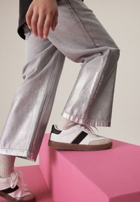 Srebrne metaliczne dżinsy wide-leg, zestawione z białymi sneakersami z czarnymi akcentami i teksturowaną brązową podeszwą, na różowej platformie.