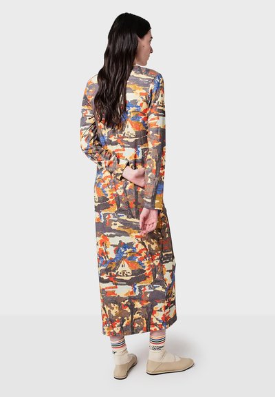 Woman with long dark hair wearing a colorful abstract-patterned midi dress, striped socks, and beige flat shoes, standing against a plain background.