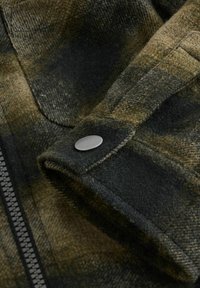 Olivgrüne karierte Flanellware mit weicher Textur, die eine Nahaufnahme eines Druckknopfs und einen sichtbaren Reißverschluss entlang der Seitennaht zeigt.