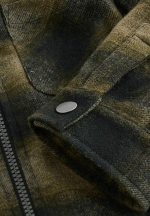Tissu flanelle à carreaux vert olive avec une texture douce, mettant en avant un gros plan d'un bouton-pression et d'une fermeture éclair visible le long de la couture latérale.