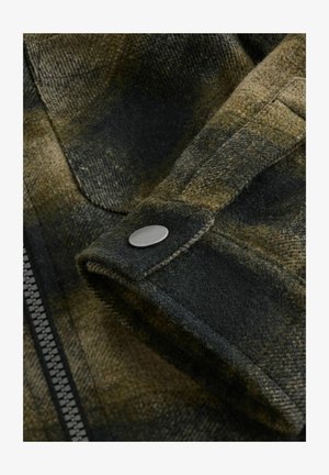 Tessuto flanella a quadri verde oliva con una texture morbida, caratterizzato da un primo piano di un bottone a pressione e una cerniera visibile lungo la cucitura laterale.