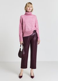 Rosa Strickrollkragenspullover mit geripptem Saum, kombiniert mit burgunderfarbenen Lederhosen. Das Model hält eine kleine dunkle Handtasche und trägt spitze High Heels.