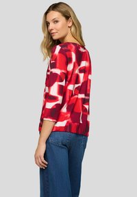 Frau mit blondem Haar, die ein Oberteil mit abstraktem Muster in Rot, Weiß und Schwarz und dreiviertellangen Ärmeln sowie blaue Jeans trägt, blickt über ihre Schulter.