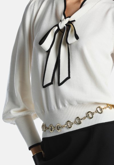 Pull blanc à manches longues avec bordure noire et nœud au niveau de l'encolure, associé à une ceinture chaine dorée et un pantalon noir.