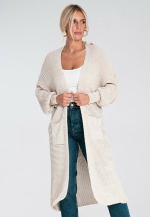 Beżowy sweter z dzianiny z otwartym przodem, długimi rękawami i dwoma przednimi kieszeniami; w zestawie z białą bluzką i niebieskimi dżinsami z wysokim stanem.