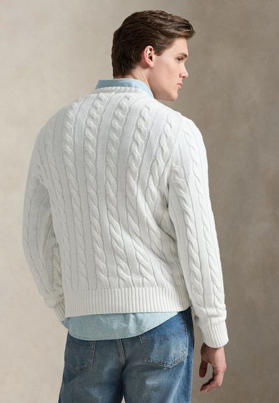 Jeune homme vu de dos portant un pull en maille torsadée blanc par-dessus une chemise bleu clair et un jean bleu, debout devant un fond neutre.