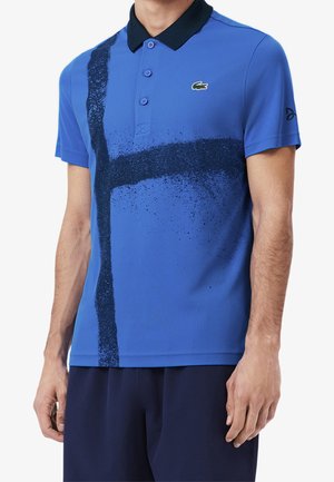 Mann trägt ein blaues Polohemd mit schwarzem Kragen und einem dunkelblauen Sprayfarben-Kreuzdesign auf der Vorderseite, kombiniert mit dunkelblauer Hose.
