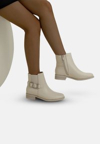 Botines beige con un acabado suave, cremallera lateral y una correa decorativa con hebilla. Tacón bajo de tipo apilado y diseño de punta redondeada.