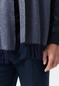 Wollschal in Dunkelblau mit Fischgrätenmuster und Fransen an den Enden. Eine Hand hält ihn vor einem strukturierten marineblauen Stoffhintergrund.