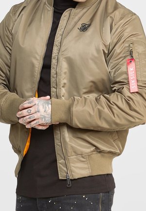 Person iført en tan bomberjakke med en rød SIKSILK-label på ærmet og hænder dækket af tatoveringer, foldet foran.