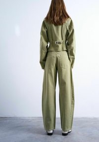 Personne aux longs cheveux bruns portant une veste vert olive et un pantalon à jambes larges, debout sur un sol en béton contre un mur blanc, vue de dos.