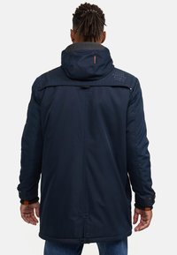Parka imperméable bleu marine avec capuche amovible, poignets ajustables et logo brodé au dos. Présente une silhouette longue et droite.