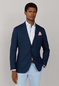 Mann trägt marineblaues Sakko, hellblau gestreiftes Hemd, hellblaue Hose und rosa gemustertes Einstecktuch, steht vor einem einfarbigen Hintergrund.