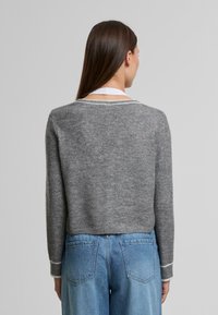 Hosszú barna hajú nő, aki szürke pulóvert és világoskék, magas derékú farmert visel, hátulról látható egy egyszínű háttér előtt.