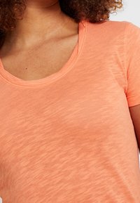 Vrouw met een kortemouwen, ronde hals oranje t-shirt, met krullend haar zichtbaar aan de bovenrand van het beeld.