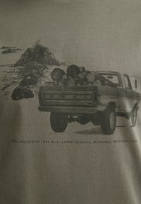 Grafische T-shirt in olijfgroen met een zwarte illustratie van een pick-up vrachtwagen vol met stammen, en tekst onderaan die het merk en de locatie aangeeft.