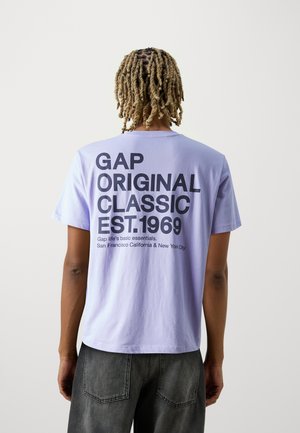Person mit blonden Dreadlocks trägt ein helllila T-Shirt mit großem schwarzem Text "GAP ORIGINAL CLASSIC EST. 1969" auf der Rückseite, kombiniert mit dunklen Jeans.