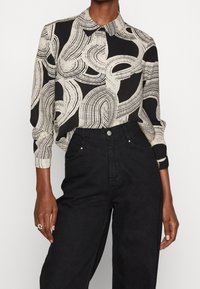 Musta ja beige kuvioitu paita, jossa on kaulus; siinä on abstrakteja viivoja ja muotoja. Yhdistetty korkeavyötäröisiin mustiin farkkuihin.