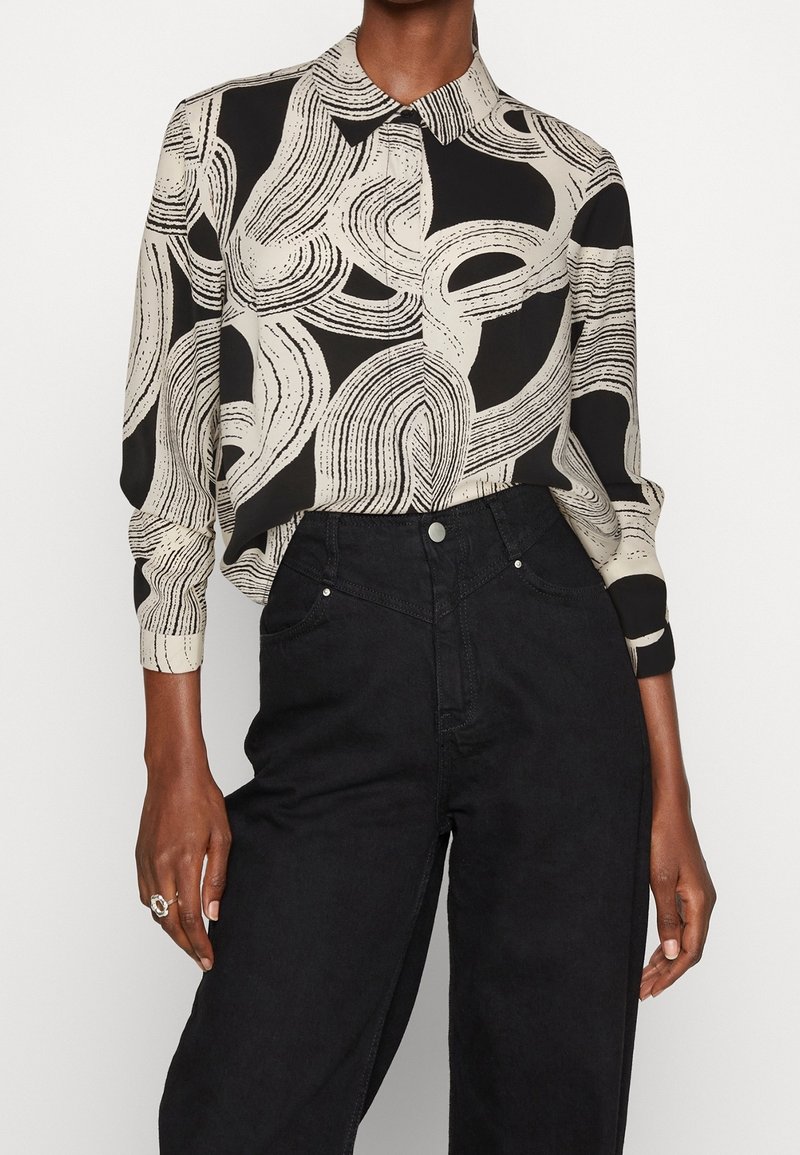 Musta ja beige kuvioitu paita, jossa on kaulus; siinä on abstrakteja viivoja ja muotoja. Yhdistetty korkeavyötäröisiin mustiin farkkuihin.