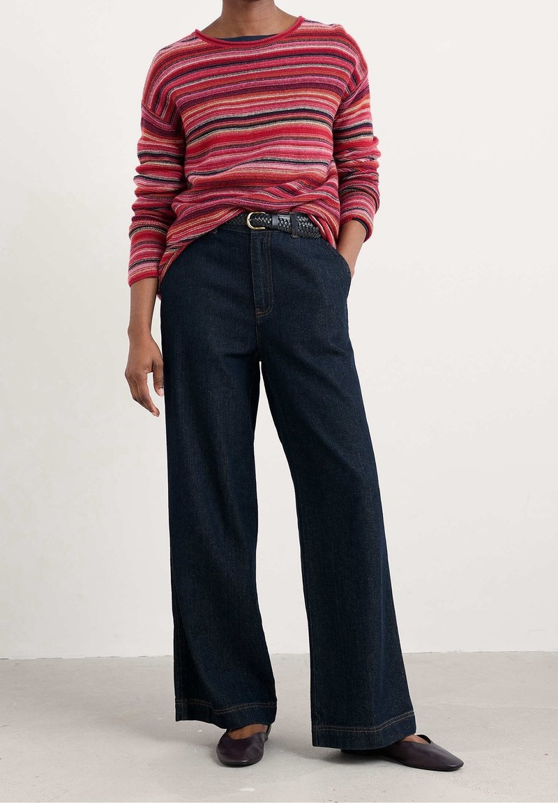 Personne portant un pull rouge à rayures à manches longues, rentré dans un jean en denim foncé taille haute à jambes larges, avec une ceinture noire et des ballerines noires.
