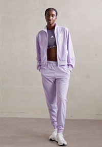 Λιλά αθλητική φόρμα που περιλαμβάνει ζακέτα με φερμουάρ και ταιριαστά παντελόνια, κατασκευασμένα από απαλό, ελαφρύ ύφασμα. Λευκά αθλητικά παπούτσια ολοκληρώνουν την εμφάνιση.