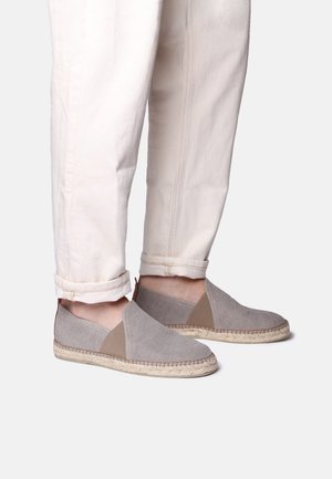 Isik, kes kannab heledate beežide pükse ja halli libisevaid espadrille-tüüpi kingi, millel on kootud joodiku tald, seisab ühevärvilise tausta ees.