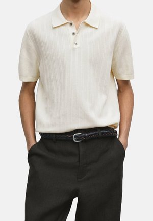 Mężczyzna ubrany w kremową, krótkorękawową dzierganą koszulkę polo z pionowymi prążkami, wpuszczoną w czarne spodnie z plecionym czarnym paskiem.