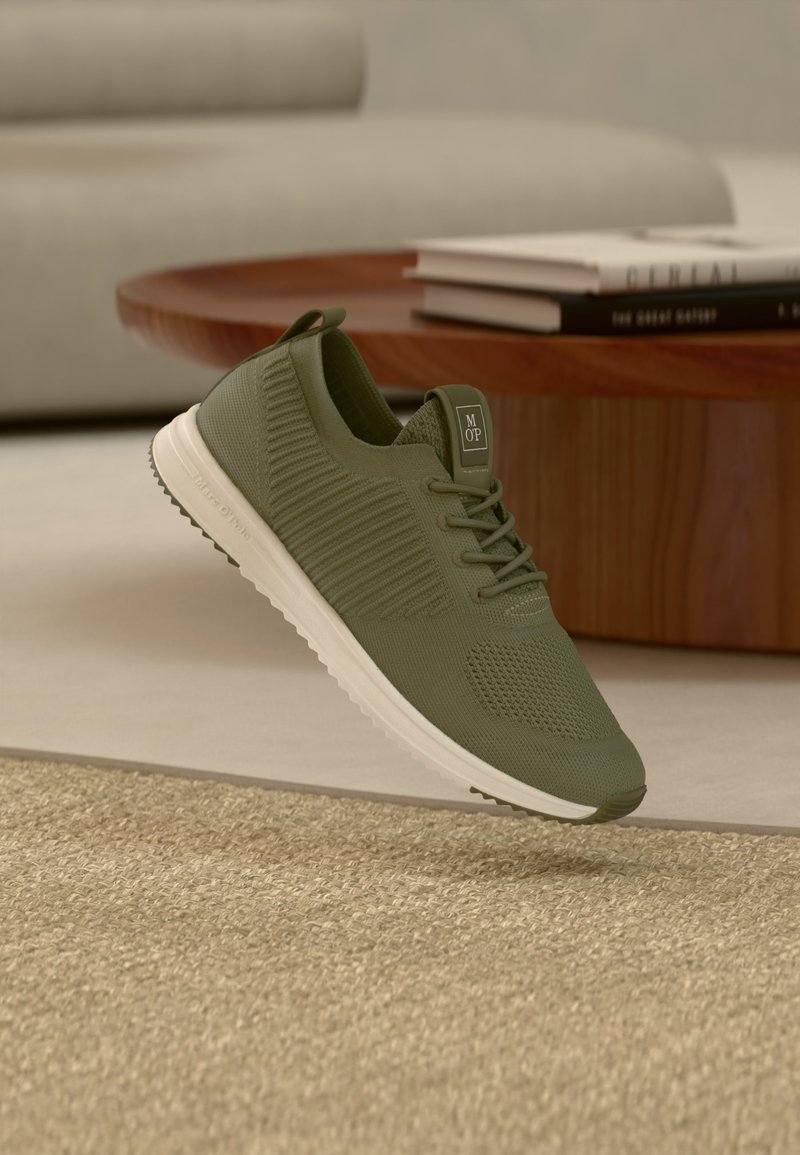Baskets en maille verte avec semelle blanche lévitant au-dessus d'un tapis beige, d'une table ronde en bois et de livres empilés dans un salon aux tons neutres.