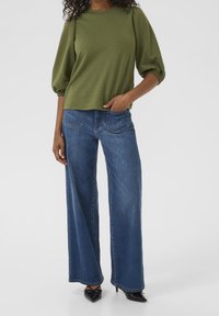 Haut vert à manches longues texturé avec épaules bouffantes ; associé à un jean large en denim bleu. Chaussures noires à bout pointu complètent la tenue.