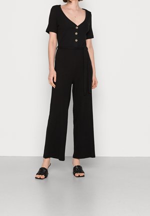 Woman wearing a black short-sleeve jumpsuit with buttons, a fabric belt, wide-leg pants, and black woven slip-on sandals.
