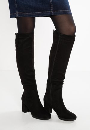 Femme portant des bottes en daim noires hauteur genou avec talons blocs, associées à des collants noirs et une jupe en jean.