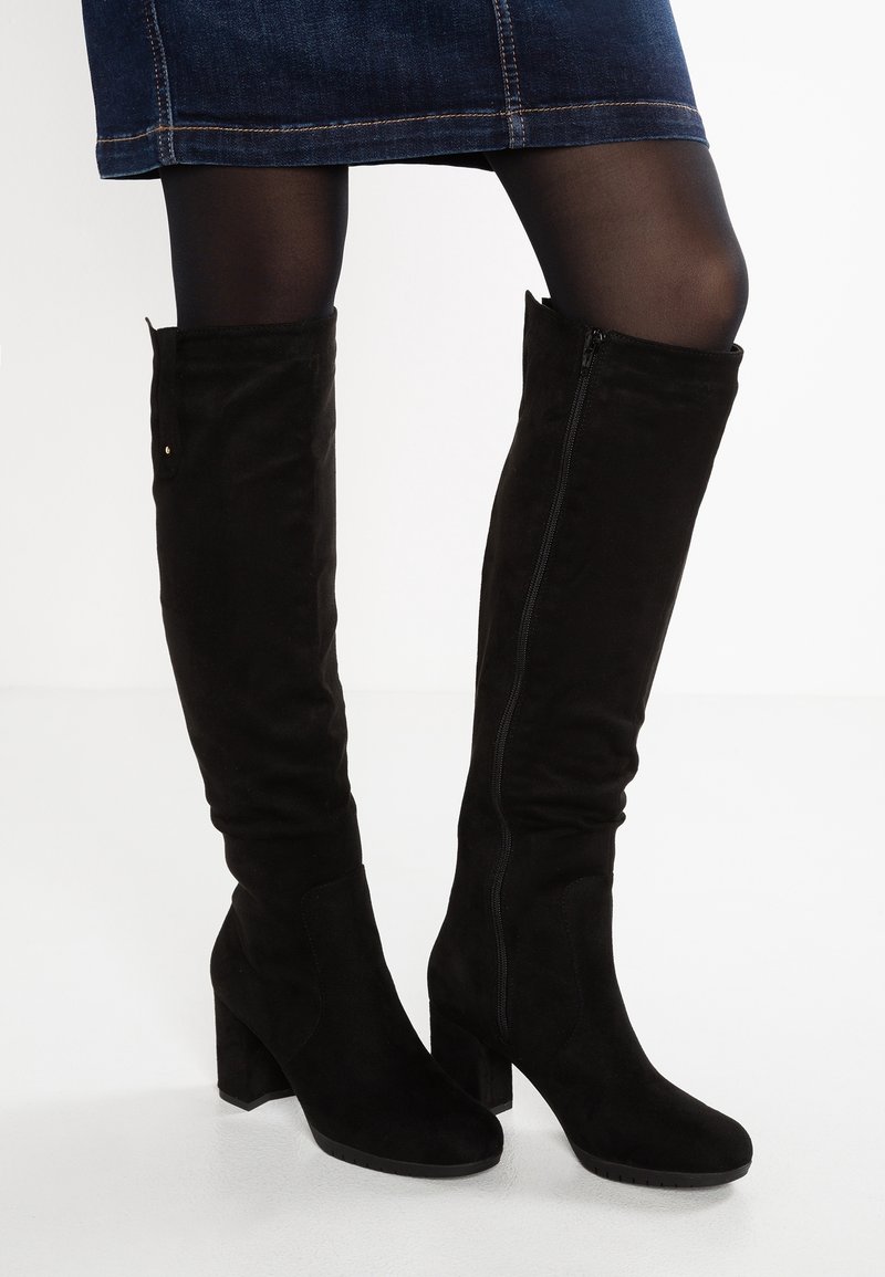 Femme portant des bottes en daim noires hauteur genou avec talons blocs, associées à des collants noirs et une jupe en jean.