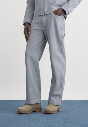 Person iført blå- og hvide pinstribede bukser med matchende jakke og brune støvler, står på et blåt tæppe.