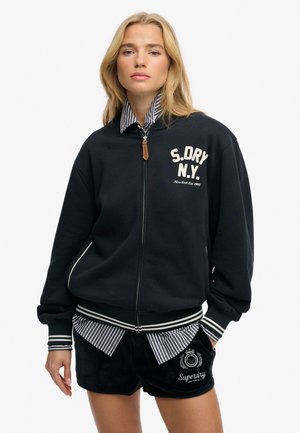 Femme blonde portant une veste bomber noire zippée avec le texte « S.DRY N.Y. », une chemise rayée en dessous et un short noir avec le logo « Superdry ».