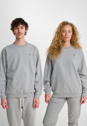 To voksne står side om side iført matchende lysegrå sweatshirts og bukser mod en simpel hvid baggrund.