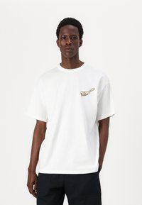 Hvid bomulds T-shirt med korte ærmer, der har et lille farverigt grafisk motiv på venstre side. Rund halsudskæring, afslappet pasform, glat tekstur.