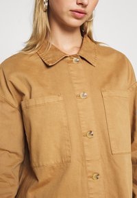 En tan knäppt skjorta tillverkad av ett tyg med en mjuk textur, med en krage, två framfickor och stora knappar.