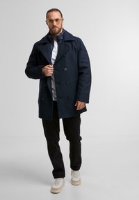 Marineblauer, doppelt geknöpfter Wollmantel mit großen Knöpfen, über einem weißen Hemd getragen, kombiniert mit schwarzen Hosen und hellen Sneakern.
