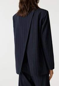 Merihenkinen pinstripe-bleiseri, jossa on halkeama selkäosassa, rakenteelliset olkapäät ja sileä tekstuuri. Muotoiltu istuvuus pitkillä hihoilla.
