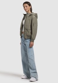 Veste bomber vert olive avec capuche, dotée de deux poches avant, d'une poche zippée sur le bras et de poignets côtelés, portée sur un jean bleu clair.
