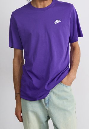 T-shirt Nike violet, manches courtes, col rond, en coton, avec un logo Nike blanc sur le côté gauche de la poitrine, associé à un jean bleu clair.