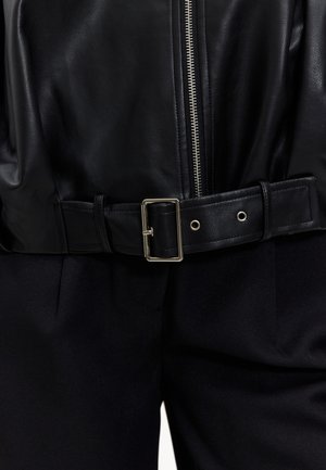 Veste en cuir noire avec une fermeture éclair argentée et une ceinture noire épaisse avec une boucle en métal. Le tissu a une texture lisse, complété par un pantalon foncé.