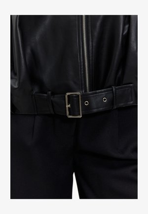 Veste en cuir noire avec une fermeture éclair argentée et une ceinture noire épaisse avec une boucle en métal. Le tissu a une texture lisse, complété par un pantalon foncé.