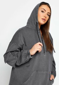 Grå huvtröja med dragkedja vid halsen, elastiska muddar och en stor framficka. Tillverkad av strukturväv med avslappnad passform.