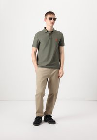 Olive zelené polo tričko s krátkým rukávem a oranžovým logem, spárované s béžovými kalhotami a černými teniskami. Model nosí tmavé sluneční brýle.